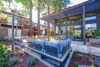 Outdoor patio with gray sectional seating, Contemporary wooden pergola providing partial shade, Floor-to-ceiling glass windows on modern home, Dining areas with minimalist tables and chairs, Surrounded by tall evergreen trees in a peaceful setting