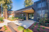 Modern building entrance with covered portico and double glass doors, landscaping features concrete patio, low-lying shrubs, groundcover, and mulch, Contrasting dark-stained wood panels and light-colored concrete