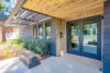 Modern building entrance with covered portico and double glass doors, landscaping features concrete patio, low-lying shrubs, groundcover, and mulch, Contrasting dark-stained wood panels and light-colored concrete