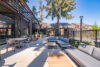 Outdoor patio with gray sectional seating, Contemporary wooden pergola providing partial shade, Floor-to-ceiling glass windows on modern home, Dining areas with minimalist tables and chairs, Surrounded by tall evergreen trees in a peaceful setting