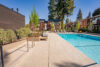 Swimming pool at apartment complex, clear blue water, deck with lounge chairs and seating, two-story buildings with cedar siding, balconies overlooking pool area, tall evergreen trees and landscaping, communal recreational space